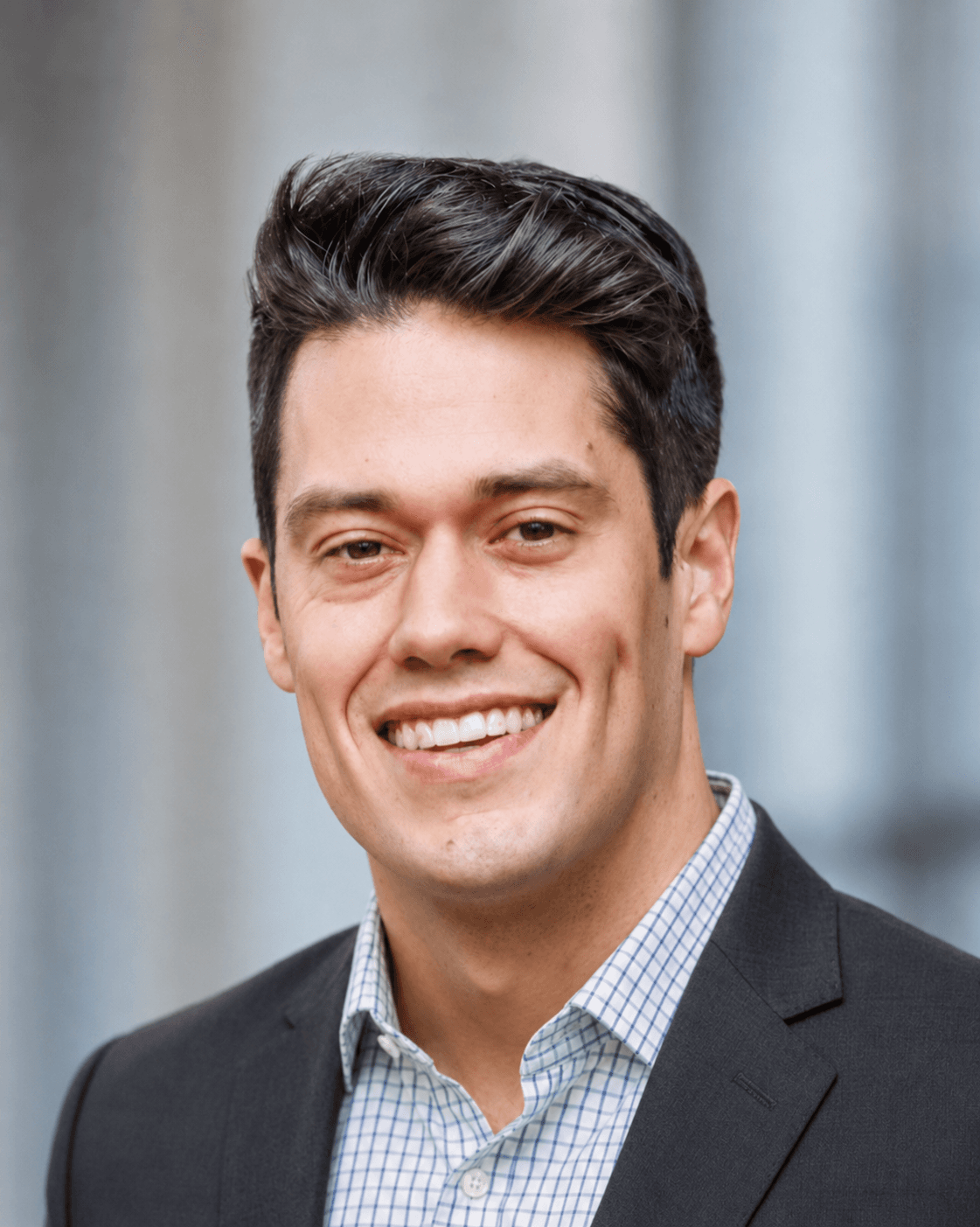 Smiling man with dark hair wearing a dark blazer and light blue checkered shirt.